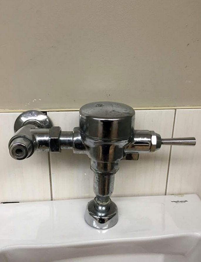 Chrome manual flush valve mounted above a fixture in a commercial restroom